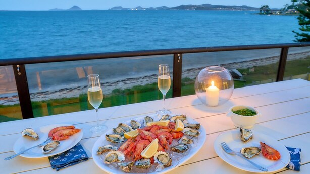 Mesa dispuesta con mariscos, incluyendo ostras y camarones, y dos copas de vino blanco, con vista al mar.