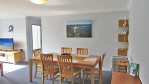 Un coin repas avec une table et des chaises en bois, un téléviseur posé sur un meuble, un tableau encadré accroché au mur et un radiateur.