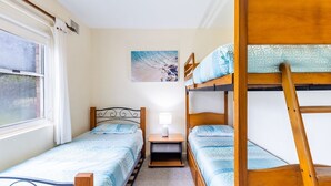 Quarto de dormitório com duas beliches, uma pequena mesa de cabeceira com abajur e uma janela com cortinas.