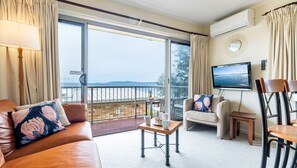 Een ruime woonkamer met een leren bank, een tafeltje en een stoel. Een televisie is aan de muur bevestigd, en er is een balkon te zien met uitzicht op zee.