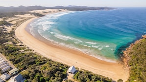 An aerial view of a coastal area with a sandy beach, a few buildings, and a clear view of the ocean. - Beach House 7,  Air Conditioned, Wi-Fi, Foxtel, Linen, Communal Pool and Spa (One Mile)