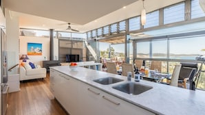 A modern kitchen with a large island, a dining area with outdoor seating, and a view of the ocean. - Beach House 7,  Air Conditioned, Wi-Fi, Foxtel, Linen, Communal Pool and Spa (One Mile)