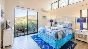 A bedroom with a large bed, a desk, a chair, and a view through a sliding door to an outdoor area. - Beach House 7,  Air Conditioned, Wi-Fi, Foxtel, Linen, Communal Pool and Spa (One Mile)