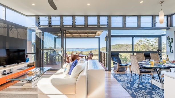 A modern living room with a large TV, sofa, and dining area with a view. - Beach House 7,  Air Conditioned, Wi-Fi, Foxtel, Linen, Communal Pool and Spa (One Mile)