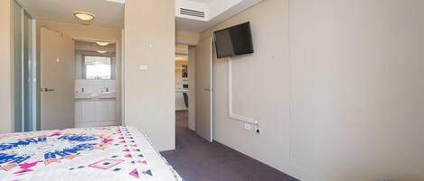 Una camera d'albergo con un letto, una TV a muro, un bagno con lavandino e specchio, e un corridoio che porta a un'altra stanza.