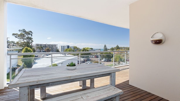 Varanda com mesa de madeira e planta em vaso, com vista para um estacionamento e edifícios.