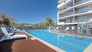 Uma piscina com espreguiçadeiras, um deck de madeira e um edifício com várias andares e varandas.