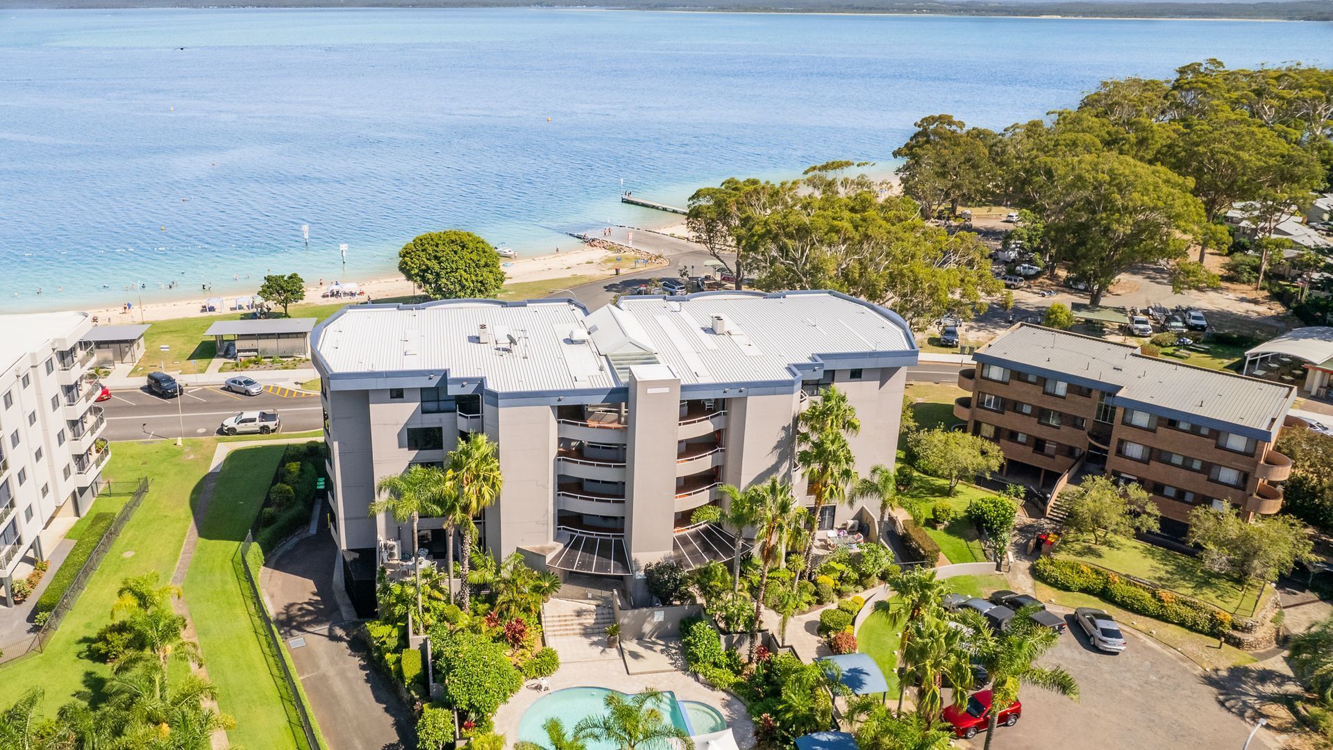 Een meerlaags hotel met een zwembad, omgeven door weelderige begroeiing en een strand.