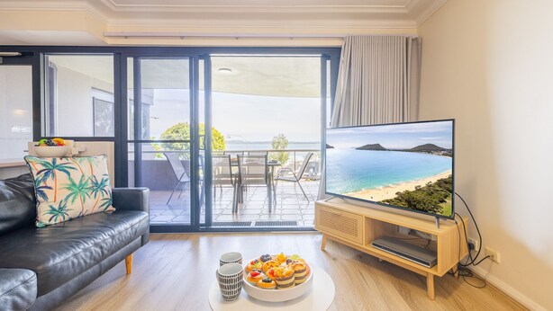 A living room with a black leather sofa, a coffee table with pastries, a TV displaying a beach view, and a balcony with a table and chairs. - Florentine, 7 11 Columbia Close - wifi, air conditioned unit with fantastic views of Little Beach (Nelson Bay)