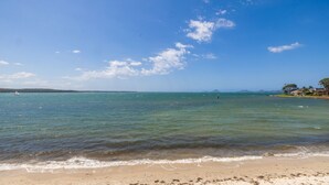 Ein Strand mit klarem Wasser, einem Sandufer und einem entfernten Blick auf Häuser und Bäume.