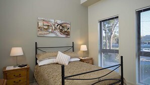 Un dormitorio con cama, dos mesitas de noche, una lámpara en cada una, una ventana con persianas y un cuadro en la pared.