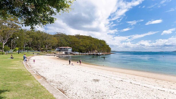 A beach with a white sandy shore, a pier extending into the water, and a building with a red roof on a grassy hill. - Collendina, 4/19 Kurrawa Close - fantastic water views and sea breezes (Nelson Bay)