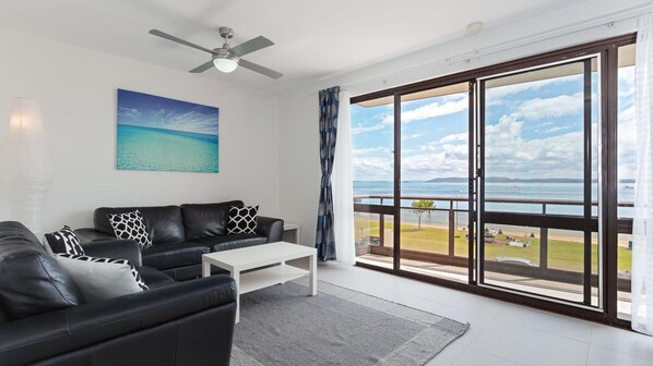 A modern living room with a black leather sofa, a white coffee table, and a painting of a beach. - Collendina, 4/19 Kurrawa Close - fantastic water views and sea breezes (Nelson Bay)