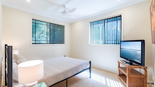 Ein Schlafzimmer mit einem Bett, einem Fernseher auf einem Holzboden, einem Deckenventilator und zwei Fenstern mit Jalousien.