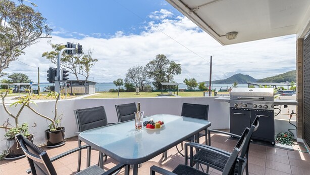 An outdoor patio with a glass table, chairs, a grill, and a view of the sea and mountains. - Fleetwood, 2/63 Shoal Bay Road - spectacular views (Shoal Bay)