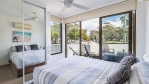 A bedroom with a large bed, a ceiling fan, and a view of the street through large windows. - Fleetwood, 2/63 Shoal Bay Road - spectacular views (Shoal Bay)