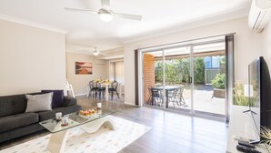 A spacious living room with a dark grey sofa, a glass coffee table, and a dining area with a brick wall and outdoor patio. - Bushmans, 9 24 Tomaree Street - Spacious townhouse (Nelson Bay)
