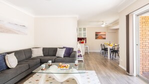 A modern living room with a grey sectional sofa, a glass coffee table, and a dining area with a white table and chairs. - Bushmans, 9 24 Tomaree Street - Spacious townhouse (Nelson Bay)