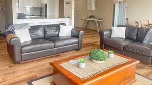 Ein modernes Wohnzimmer mit zwei grauen Ledersofas, einem Couchtisch mit Pflanzen und einer Küche im Hintergrund.