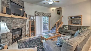 A living room with a stone fireplace, a television, a ceiling fan, a sofa, and a staircase.
