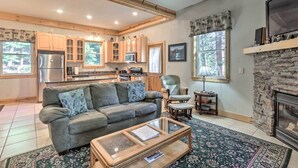 A spacious living area with a sofa, coffee table, and fireplace. A kitchen with wooden cabinets and a stone fireplace wall is visible in the background.