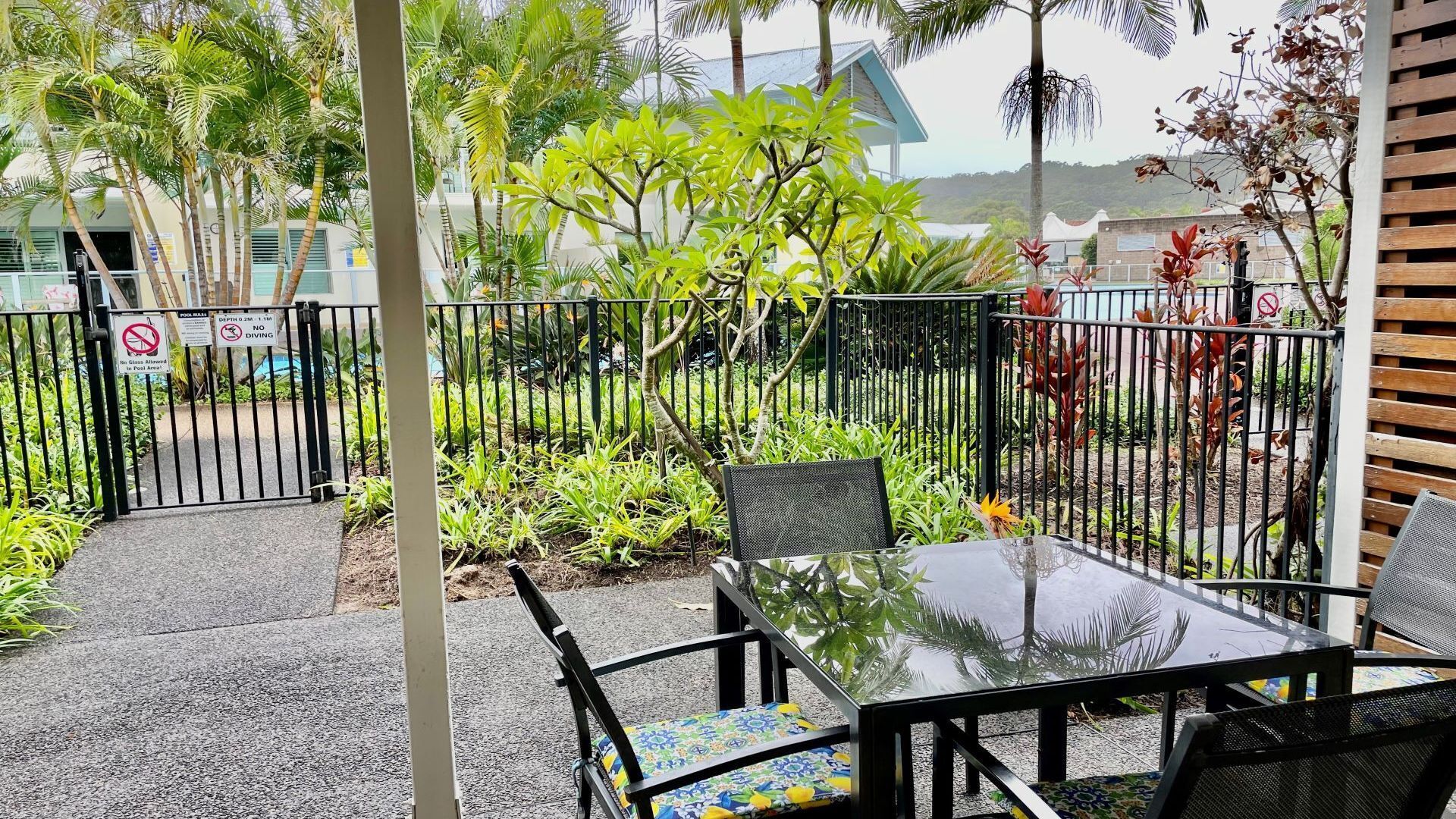 An outdoor seating area with a table and chairs, surrounded by a metal fence and lush greenery.