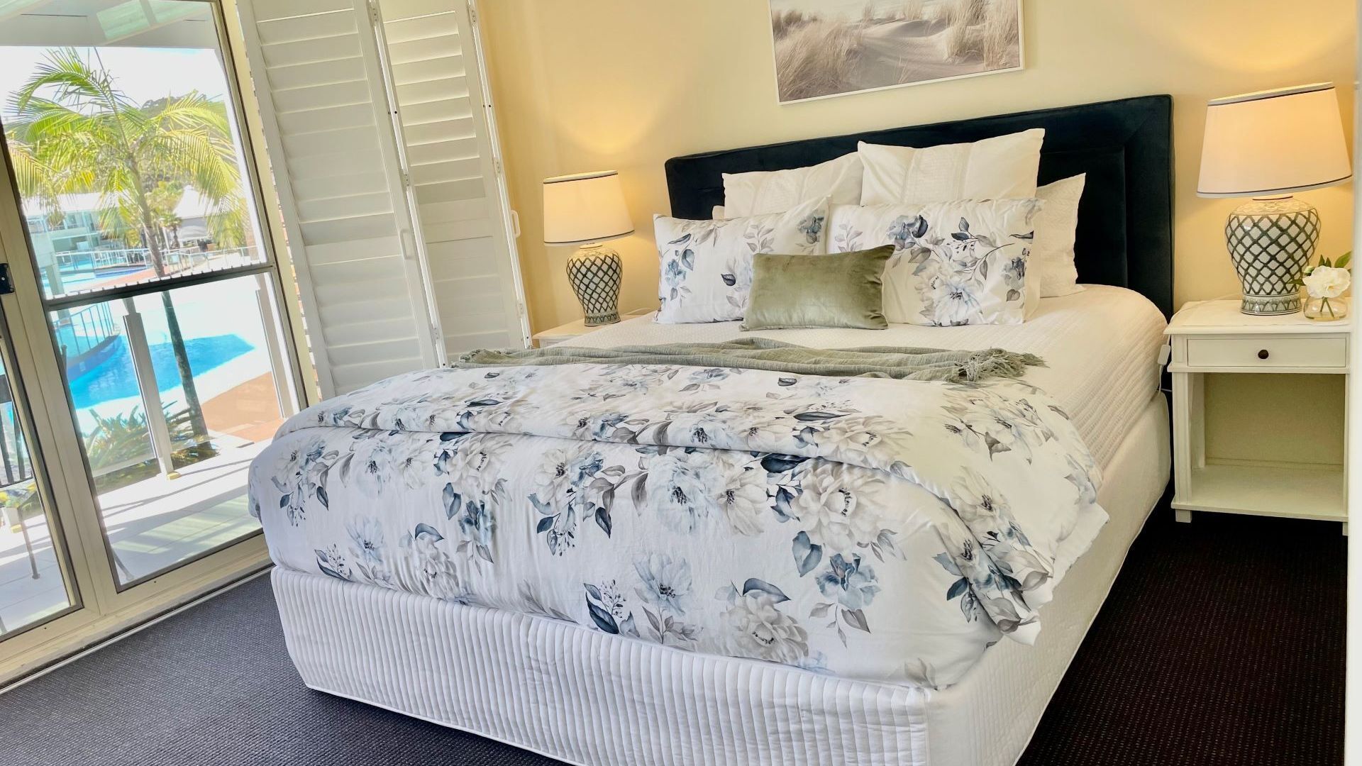A hotel room with a large bed, a nightstand, a lamp, and a view of a pool through a sliding door.
