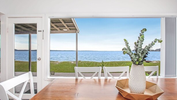 A dining area with a view of the sea, white chairs, and a wooden table. - Beached Inn, 93 Foreshore Drive - Spacious beach front house (Salamander Bay)