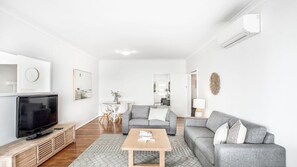 A modern living room with a flat-screen TV, grey sofas, a wooden coffee table, and a white wall with minimal decor.