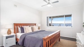 A bedroom with a wooden bed, white walls, a window with a view of the sea, a ceiling fan, and a nightstand with a lamp.