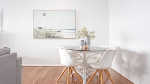 A dining area with a round glass table, white chairs, and a framed picture of a lighthouse on the wall.