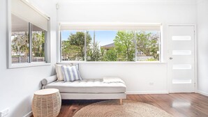 A modern living room with a grey sofa, a round wicker side table, a large window with a view of greenery and buildings, and a white door. - Beached Inn, 93 Foreshore Drive - Spacious beach front house (Salamander Bay)