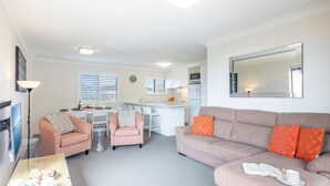 A modern living room with a sofa, armchairs, and a coffee table. There is a kitchen in the background with a dining area. - Intrepid, 6/3 Intrepid Close - right on the waterfront (Nelson Bay)