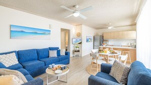 A modern living room with a blue sofa set, a coffee table with a meal, and a kitchen area with wooden cabinets and a dining table. - Shoal Towers, unit 1 11 Shoal Bay Road -  fantastic unit across the road from beach with Wi-Fi (Shoal Bay)