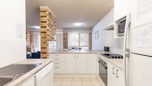 Een moderne keuken met witte kastjes, een ingebouwde oven en een magnetron. In het midden staat een bakstenen zuil, en op de achtergrond een raam met jaloezieën.