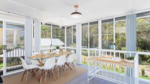 Een zonnige kamer met een glazen tafel omringd door witte stoelen, grote ramen met uitzicht op groen, en een hanglamp.