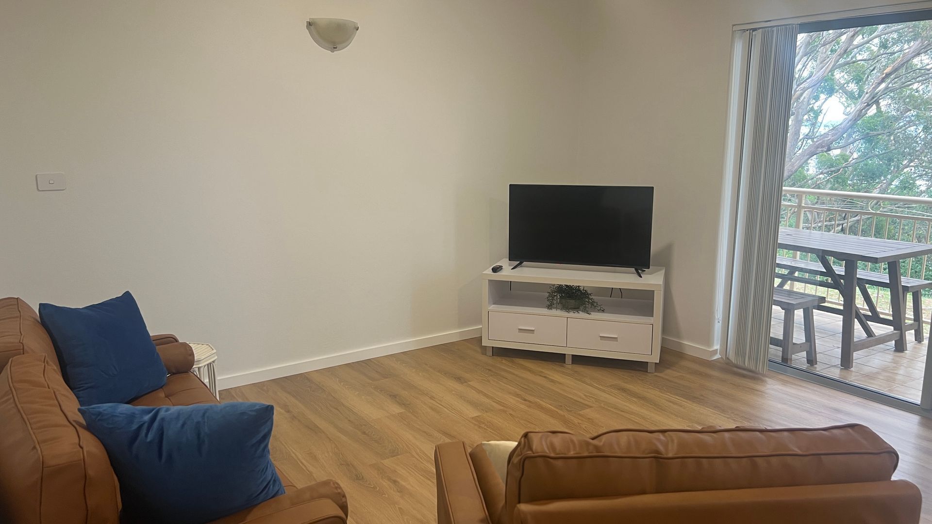 Una sala de estar con un sofá de cuero marrón, un mueble blanco para televisión con una pantalla plana y un ventanal corredizo que da a un patio exterior.