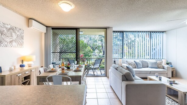 Una sala moderna con sofá, mesa de comedor y sillas. Hay un balcón con vistas a zonas verdes y una ventana con persianas.