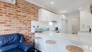 A modern kitchen with white cabinets, a brick wall, and a leather couch. - Bay Parklands, 68 2 Gowrie Ave - Fully ducted aircon, wifi, pool, tennis court, communal spa (Nelson Bay)