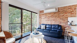 A modern living room with a blue leather sofa, a round white table, and a brick wall. - Bay Parklands, 68 2 Gowrie Ave - Fully ducted aircon, wifi, pool, tennis court, communal spa (Nelson Bay)