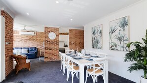 A dining area with a brick wall, a blue sofa, white chairs, and a table set for four. - Bay Parklands, 68 2 Gowrie Ave - Fully ducted aircon, wifi, pool, tennis court, communal spa (Nelson Bay)