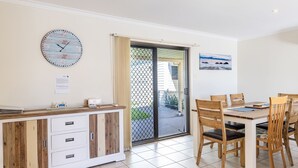 A dining area with a wooden table and chairs, a clock on the wall, and a sliding glass door leading outside. - Happy Tails , 55 Pantowora Street - pet friendly, air conditioned (Corlette)