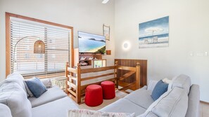 A living room with a TV, wooden furniture, red stools, and a painting of a beach scene. - Plattens, 2/15 Kurrawa Close - great duplex so close to the water (Nelson Bay)