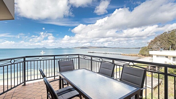 A balcony with a table and chairs, overlooking a calm sea with a pier and distant land. - 2/137 Soldiers Point Road - luxury unit on the waterfront with aircon and free unlimited WiFi (Soldiers Point)