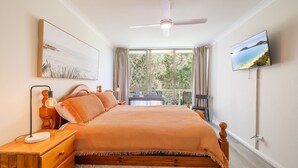 A bedroom with a bed, a desk, a chair, a ceiling fan, a TV, and a window with a view of greenery. - Mistral Court, 14/17 Mistral Close - across the road from the beach (Nelson Bay)