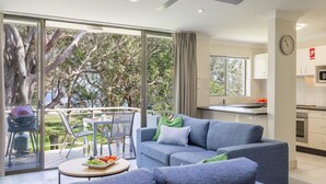 A modern living room with a grey sofa, a dining table with a fruit platter, and a view of trees through large glass doors. - Mistral Court, 14/17 Mistral Close - across the road from the beach (Nelson Bay)