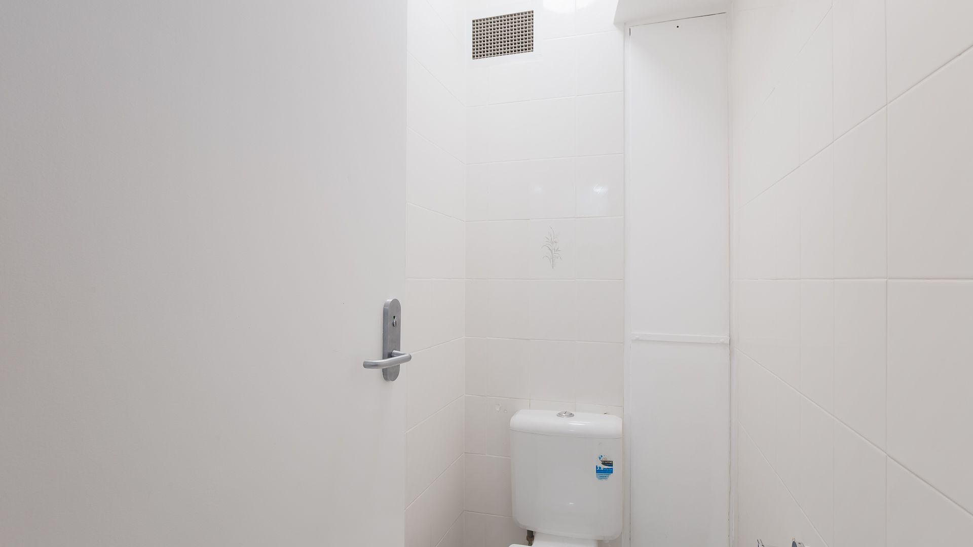 Et badeværelse med hvide fliser, et toilet og en dør med greb.