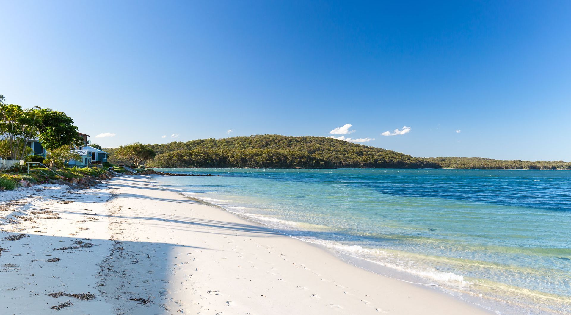 A beach with clear turquoise water, white sand, and a forested hill in the background.