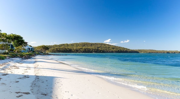A beach with clear turquoise water, white sand, and a forested hill in the background.