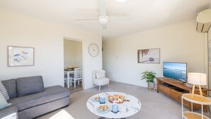 A modern living room with a grey sofa, a round coffee table with snacks, a TV, and a ceiling fan. - Yarramundi, 4,47 Magnus Street - air conditioned unit with air con, wi-fi, water views and close to town (Nelson Bay)